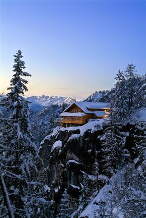 Die Dolomitenhütte liegt eingebettet in eine Winterlandschaft auf dem Felsen. Im Hintergrund färbt sich der Himmel leicht und Abendstimmung kommt auf.