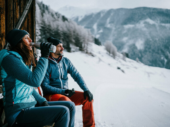 Beim Winterwandern in Kals am Großglockner in Osttirol trinkt eine Frau aus einer Flasche, ihr Partner sitzt daneben und sieht in die verschneite Landschaft