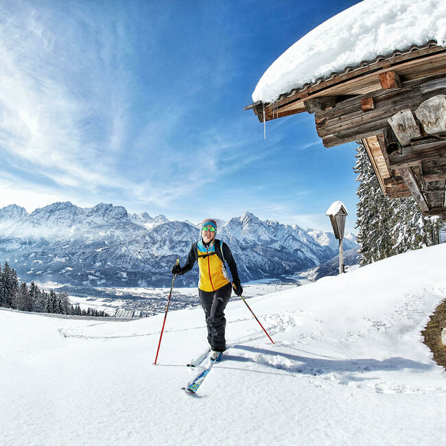 Eine Skitourengeherin neben einer kleinen, verschneiten Holzhütte am Zettersfeld mit Lienzer Talboden im Hintergrund.