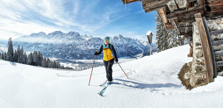 Eine Skitourengeherin neben einer kleinen, verschneiten Holzhütte am Zettersfeld mit Lienzer Talboden im Hintergrund.