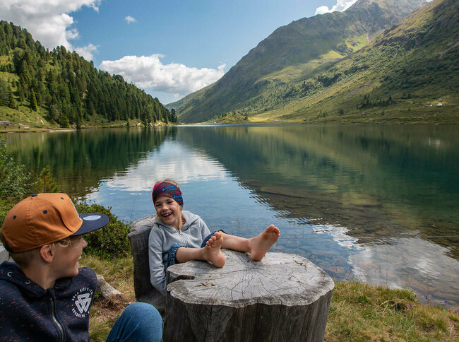 Zwei lachende Buben, die am Rand eines Gebirgssees sitzen, wobei einer seine bloßfüßigen Beine auf einem glatten, aber verwitterten Baumstumpf liegen lässt. Im Hintergrund spiegelt sich der Horziont im glasklaren Wasser des Gebirgssees.