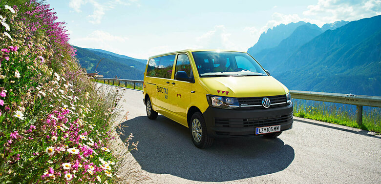 Ein gelber VW Bus fährt auf einer Straße, rechts sieht man eine Böschung mit blühenden Sommerblumen