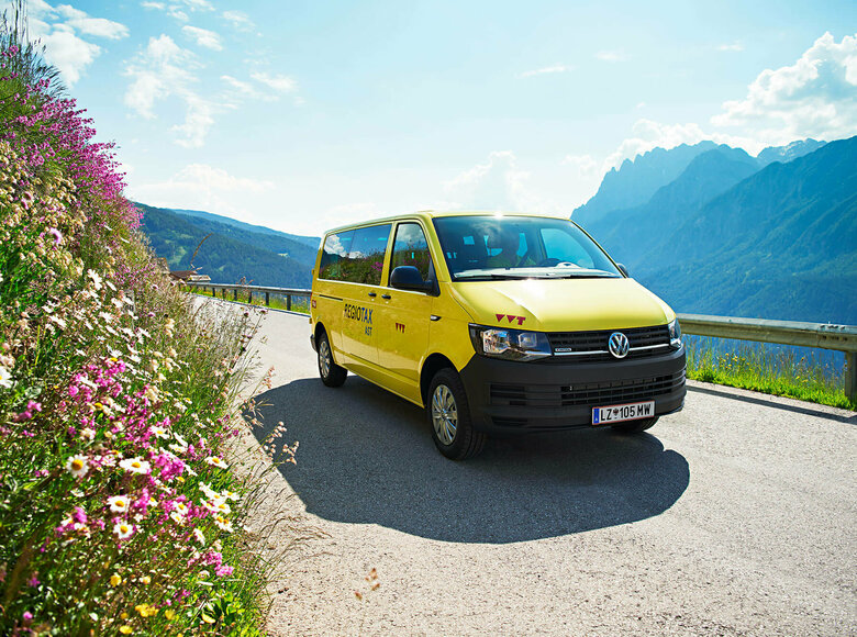 Ein gelber VW Bus fährt auf einer Straße, rechts sieht man eine Böschung mit blühenden Sommerblumen