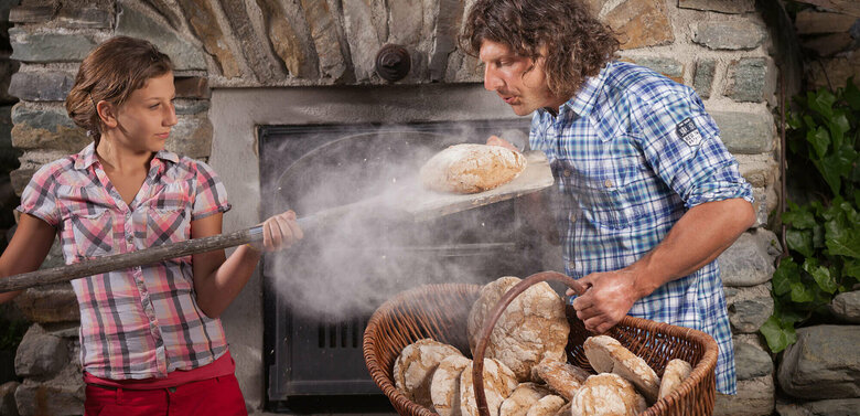 Ein Mann und eine Frau beim Brot backen. Die Frau holt ein frisch gebackenes Brot aus dem Ofen. Der Mann hält einen Korb mit mehreren Laib Brot.
