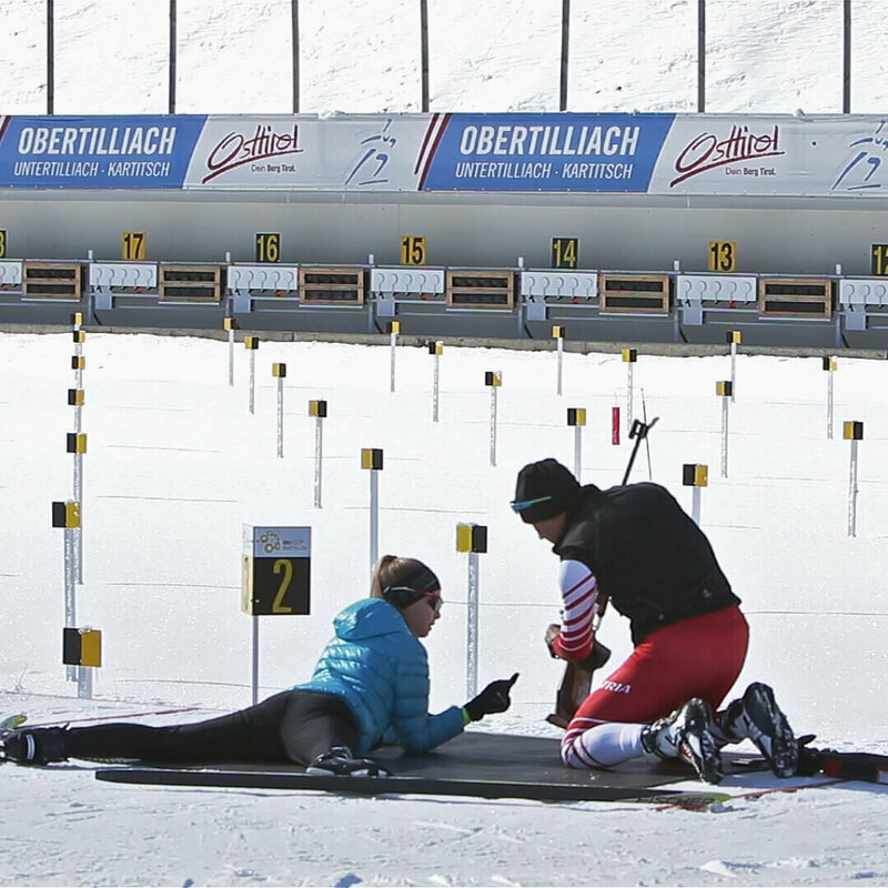Eine Frau versucht sich am Schießstand im Biathlonzentrum Obertilliach am Schießen. Dabei wird sie von einer rechts neben ihr knieenden Person unterstützt.