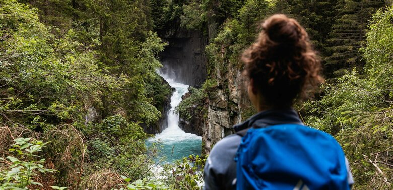 Eine Frau mit blauem Rucksack und braunem Dutt steht vor einem Wasserfall mit Blickrichtung zum Wasser.
