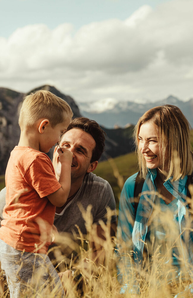Zu sehen ist ein Elternpaar mit ihrem kleinen Sohn, die im hohen Gras entlang der Kindermeile am Golzentipp den Moment zu dritt genießen.
