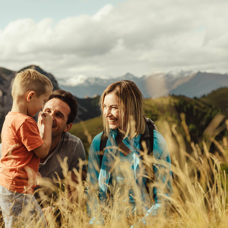 Zu sehen ist ein Elternpaar mit ihrem kleinen Sohn, die im hohen Gras entlang der Kindermeile am Golzentipp den Moment zu dritt genießen.