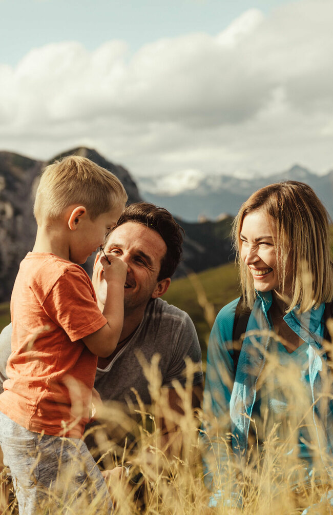Zu sehen ist ein Elternpaar mit ihrem kleinen Sohn, die im hohen Gras entlang der Kindermeile am Golzentipp den Moment zu dritt genießen.