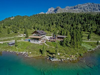 Das Alpengasthaus Obersee im Sommer mit dem darunterliegenden blau-grünen Seeufer am Staller Sattel. Im Hintergrund ist der dichte grüne Wald, der strahlendblaue Himmel und das beeindruckende Bergpanorama zu sehen.