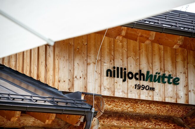 Die Aufnahme zeigt einen Ausschnitt der Niljochhütte in Virgen mit ihrer Holzfassade und dem dunklen Dach. Auf der Fassade ist gut sichtbar der Schriftzug „nilijochhütte“ sowie die Höhenangabe „1990 m“ angebracht. Die warme Holzoberfläche und die traditionelle Bauweise unterstreichen den alpinen Charakter der Berghütte.Geben Sie Ihr Feedback zu BizChat ab.