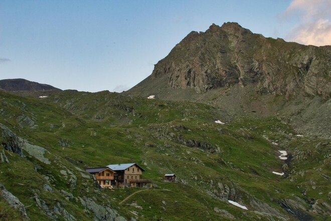 Das Bild zeigt die Eisseehütte in einer hochalpinen Berglandschaft. Die Hütte liegt auf einem grünen Berghang unterhalb eines markanten Felsgipfels und ist von Wiesen, Felsen und einzelnen Schneefeldern umgeben. Ein Wanderweg führt durch die Landschaft, während sich darüber der steile Berghang bis zum Gipfel erhebt.