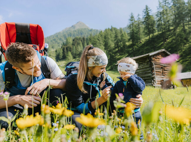 Eine kleine Familie ist im Zuge einer Familienwanderung auf einer Wiese im Kristeinertal. Die Frau und das Kind tragen Stirnbänder und der Mann hat einen roten Trage-rucksack auf dem Rücken. Sie sitzen im grünen Gras.