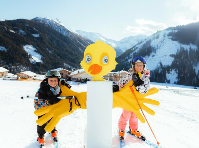 Zwei Kinder stehen im familienfreundlichen Skigebiet Golzentipp in Obertilliach hinter einem gelben Kurvenzeiger in Entendesign mit winterlichem Bergpanorama im Hintergrund.