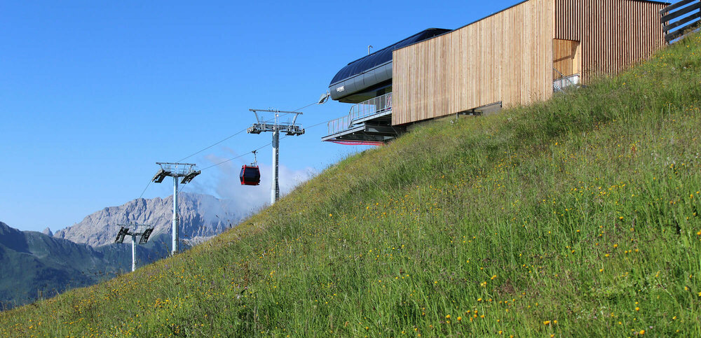 Blumenreiche, sattgrüne Bergwiese mit Seilbahn-Bergstation in senkrecht verlaufender, leicht verwitterter Holzverkleidung. Die letzten drei Liftstützen vor der Bergstation und eine rote Seilbahngondel sowie der blaue Himmel und im Hintergrund eine halbrunde, markante Felsburg bilden einen schönen Kontrast dazu.