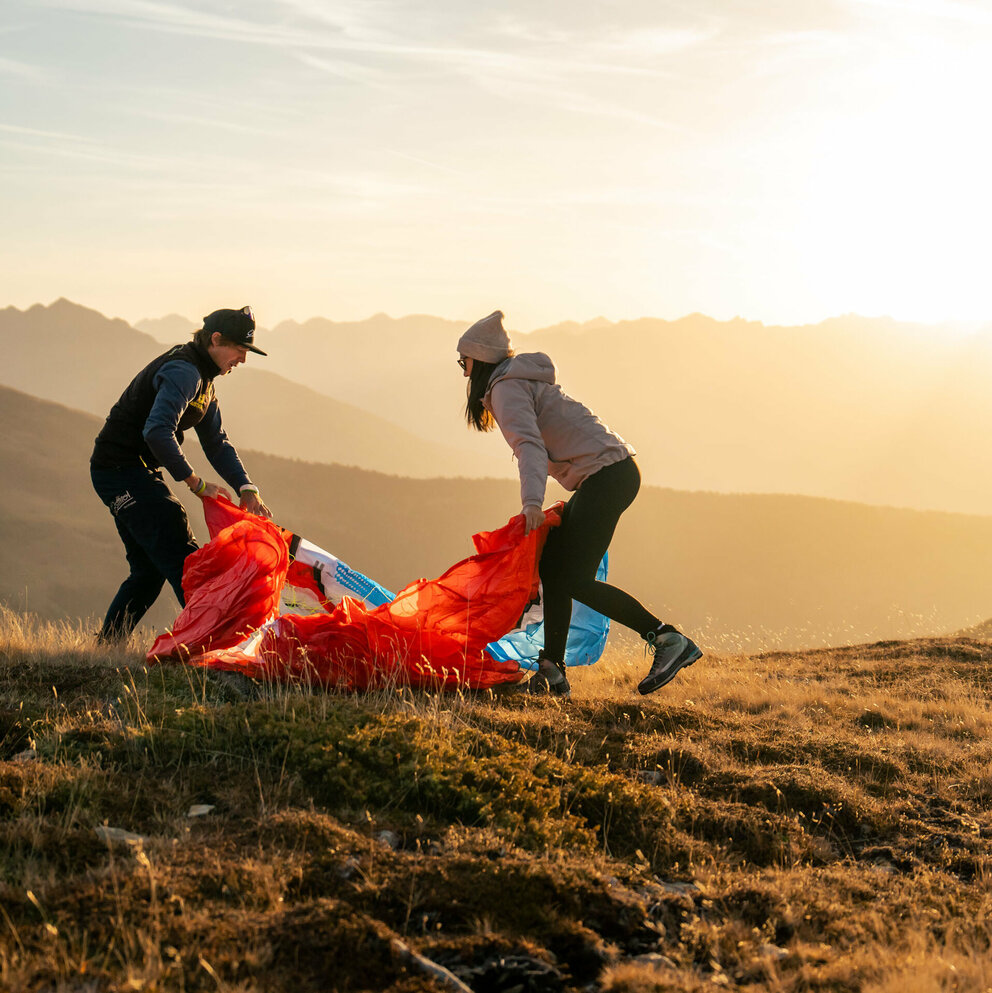 Ein Mann und eine Frau stehen auf einer Bergwiese in Osttirol in der Morgensonne und falten einen roten Gleitschirm zusammen.