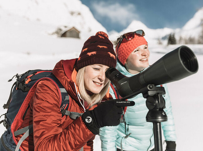Zwei Frauen bei der Schneeschuhwanderung aus der Nähe, eine blickt durch ein Fernglas 