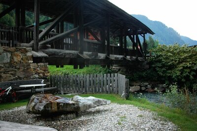 Der Wasserruheplatz bei der Blosbrücke in Hopfgarten in Defereggen in den Sommermonaten. Das grüne Gras bedeckt das Ufer neben der blauen Schwarzach, über welche die robuste Holzbrücke ragt.