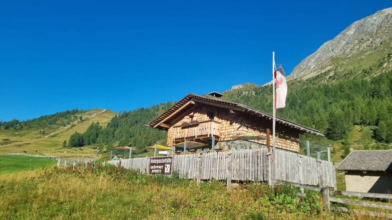 Holzhütte mit Schindelfassade und Holzzaun vor bewaldeten Berghängen unter blauem Himmel.