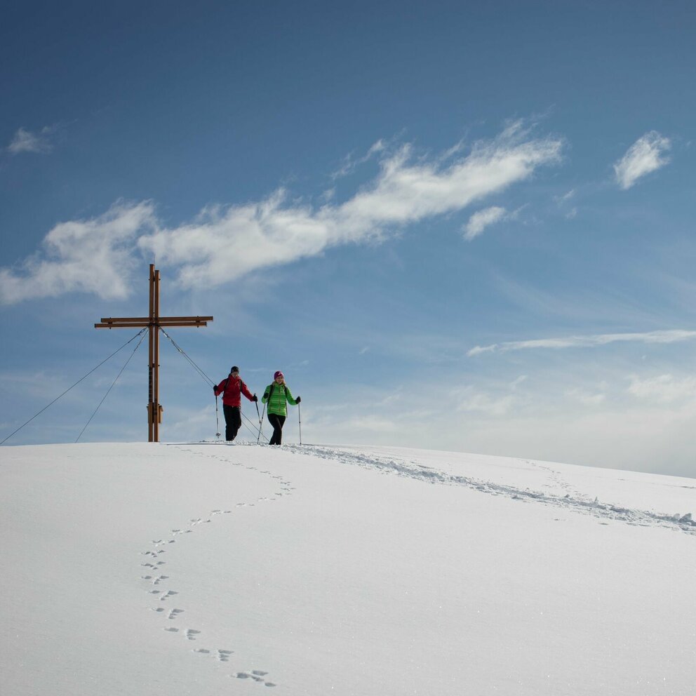 Zwei Personen stehen beim Gipfelkreuz des Dorfbergs umgeben von einer bezaubernden Winterlandschaft. Im Schnee sind außerdem Tierspuren zu erkennen.