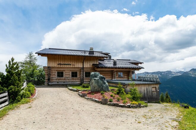 Die Nilljochhütte in Virgen ist eine Holzberghütte mit Satteldach in erhöhter Lage. Vor der Hütte verläuft ein heller Kiesweg, daneben befinden sich ein Blumenbeet und ein großer Stein. Im Hintergrund sind bewaldete Berghänge, ein Tal sowie ein Himmel mit großen weißen Wolken zu sehen.
