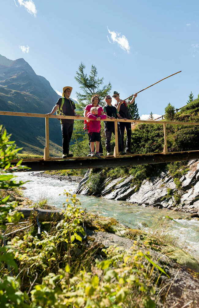 Ein Ranger zeigt einer Familie den Nationalpark Hohe Tauern bei einer geführten Rangertour. Die Gruppe steht auf einer hölzernen Brücke über einem Fluss und im Hintergrund ragt eine Bergkette empor.