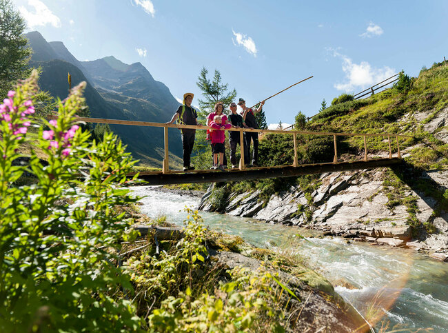 Ein Ranger zeigt einer Familie den Nationalpark Hohe Tauern bei einer geführten Rangertour. Die Gruppe steht auf einer hölzernen Brücke über einem Fluss und im Hintergrund ragt eine Bergkette empor.