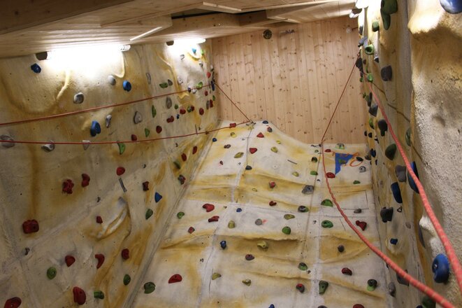 Das Bild zeigt den Kletterturm in der Sajathütte. Die Kletterwände sind mit zahlreichen bunten Klettergriffen ausgestattet und durch mehrere Kletterseile gesichert. Die Wände reichen bis unter die Holzdecke, an der Rückwand sind Holzverkleidungen und eine Lampe zu sehen.