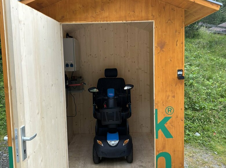 Eine hölzerne Hütte mit geöffneter Tür. Im Inneren steht der E-Scooter für barrierefreies Entdecken des Nationalparks Hohe Tauern.