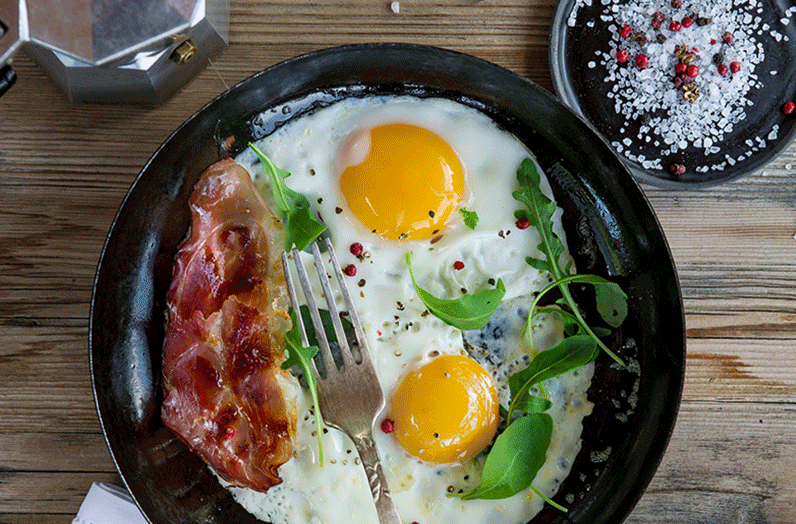 Zwei gebratenen Spiegeleier mit Schnittlauch und einer gebratenen Speckscheibe in einer Eisenpfanne mit frischen Tomaten auf der Rispe davor auf einem weißen Tuch schön angerichtet.