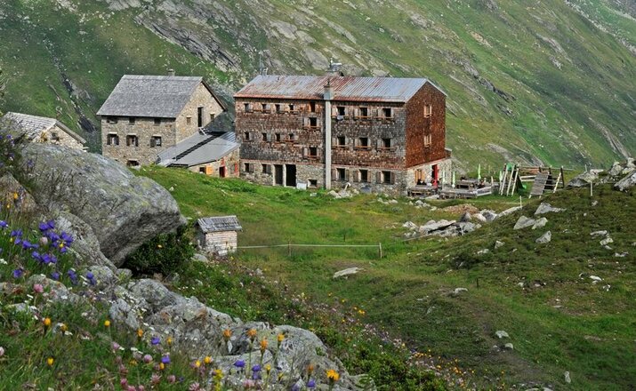 Das Bild zeigt die Essener-Rostocker Hütte mit einem mehrstöckigen Holzgebäude und einem angebauten Steingebäude. Im Vordergrund sind eine Bergwiese mit bunten Blumen, Felsen und einzelne Steine zu sehen. Rechts neben den Gebäuden befinden sich Sitzmöglichkeiten und ein kleiner Kinderspielplatz, dahinter erstreckt sich ein grüner Berghang.