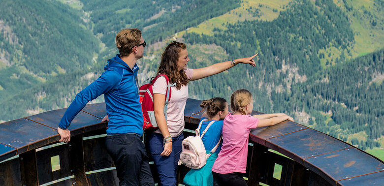 Eine vierköpfige Familie genießt den Ausblick bei der Aussichtsplattform des Wassermythos Ochsenlacke im Skizentrum St. Jakob i. D.. Ihr Blick fällt auf das gegenüberliegende Bergpanorama des Defereggentals, welches trotz einiger Wolken am Himmel in der Sonne erstrahlt.