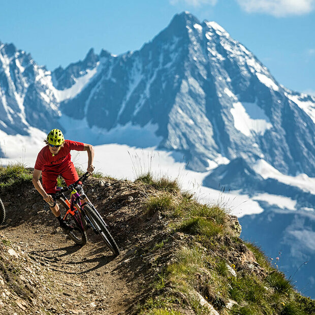 Zwei Radfahrer mit roten Shirts und gelben Helmen auf einem schmalen Trail im Bikepark Grossglockner Resort. Im Hintergrund ragt der Großglockner in den blauen Himmel.