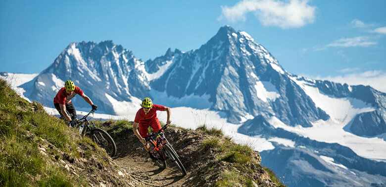 Zwei Radfahrer mit roten Shirts und gelben Helmen auf einem schmalen Trail im Bikepark Grossglockner Resort. Im Hintergrund ragt der Großglockner in den blauen Himmel.