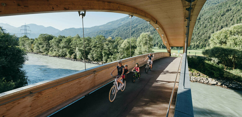 Die Abbildung zeit eine Familie, die mit ihren Fahrrädern auf dem Drauradweg unterwegs sind, und gerade eine hölzerne Brücke überqueren.