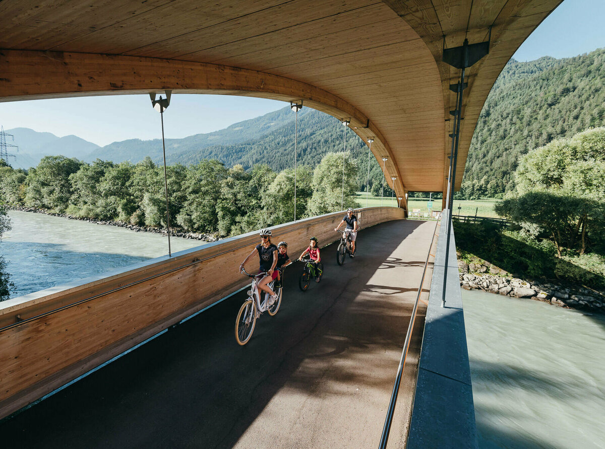 Die Abbildung zeit eine Familie, die mit ihren Fahrrädern auf dem Drauradweg unterwegs sind, und gerade eine hölzerne Brücke überqueren.