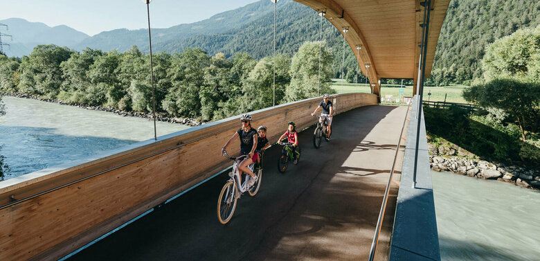 Die Abbildung zeit eine Familie, die mit ihren Fahrrädern auf dem Drauradweg unterwegs sind, und gerade eine hölzerne Brücke überqueren.