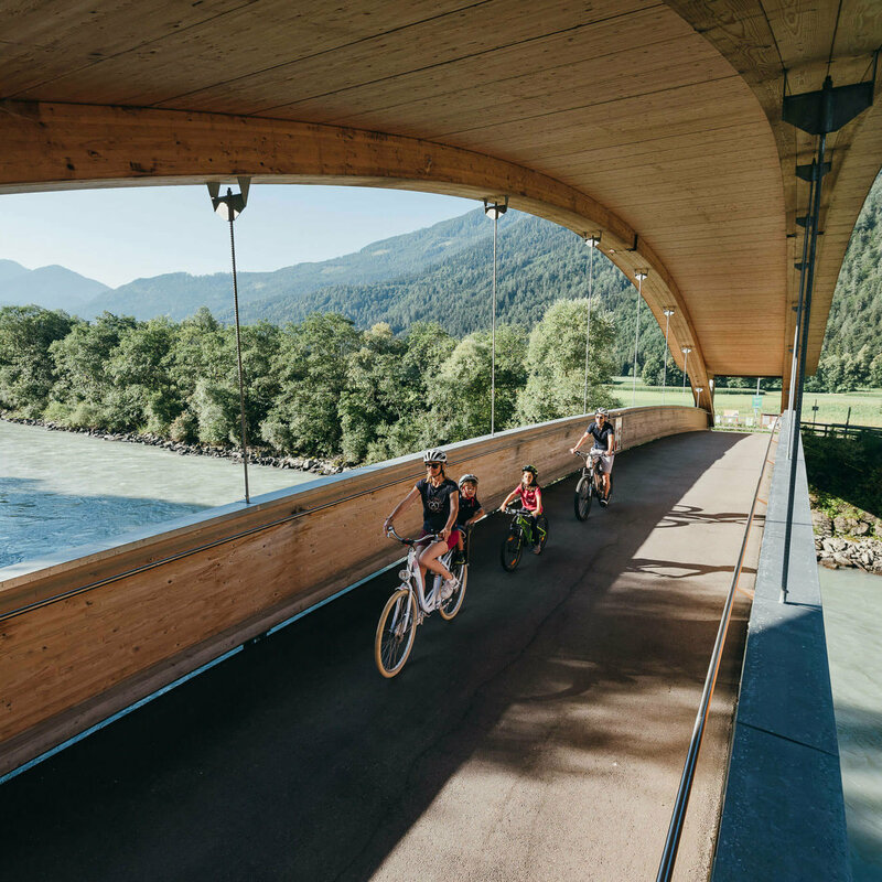 Die Abbildung zeit eine Familie, die mit ihren Fahrrädern auf dem Drauradweg unterwegs sind, und gerade eine hölzerne Brücke überqueren.