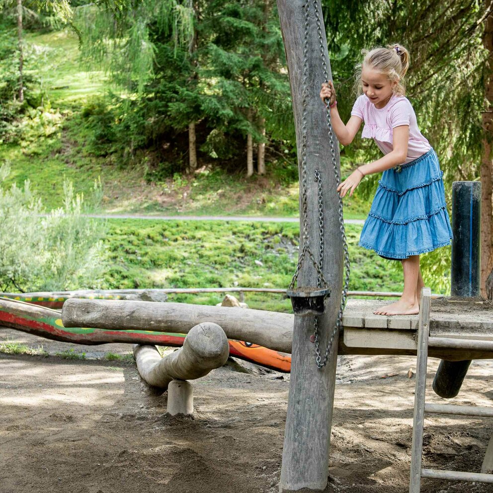 Kinder spielen auf dem Wassernaturspielplatz in St. Jakob i. D. Sie klettern barfuß auf den hölzernen Spielgeräten, welche auf einem sandigen Untergrund stehen. Im Hintergrund ist der grüne dichte Wald zu sehen, welcher dem Spielplatz genügend Schatten für den sonnigen Tag spendet.