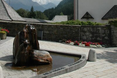 Der Nepomuk-Brunnen im Zentrum von Hopfgarten i. D., direkt neben der Pfarrkirche gelegen. Das Wasser sprudelt aus dem mehrfärbigen Stein. Im Hintergrund ist die Kirchenmauer in die davorliegende Blumenpracht an einem sonnigen Sommertag zu sehen.