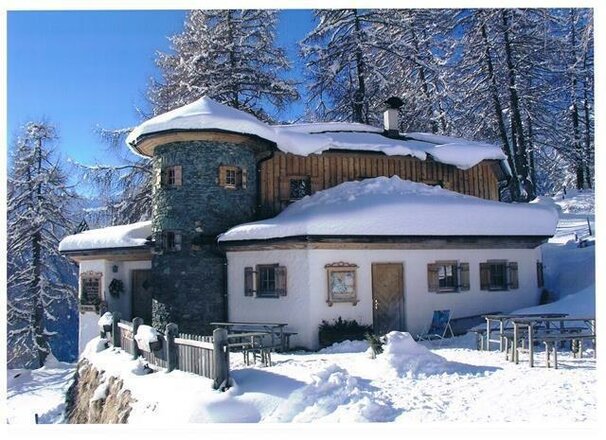 Das Bild zeigt die Stabanthütte im Winter, umgeben von einer dicken Schneedecke. Das zweigeschossige Gebäude mit heller Fassade, Natursteinmauerwerk und Holzverkleidung hebt sich deutlich vor den verschneiten Bäumen im Hintergrund ab. Vor der Hütte sind die teilweise eingeschneiten Sitzgelegenheiten und der verschneite Zugangsbereich zu sehen.