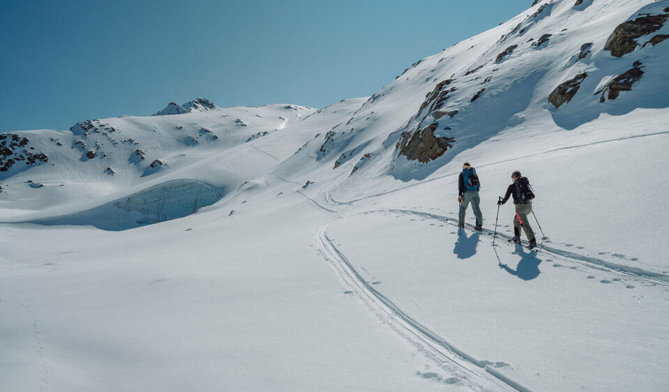 Zwei Menschen beim Skitourengehen, hinterlassen Spuren im unberührten Schnee, umgeben von schneebedeckten Bergen unter klarem Himme