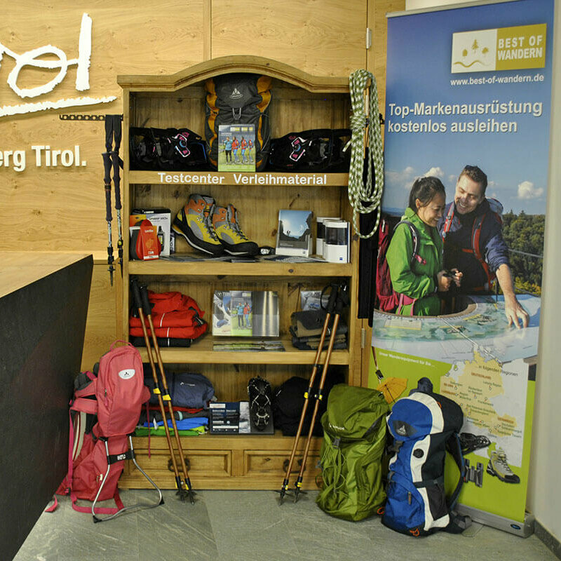 Ein Regal voller Wander-Ausrüstung. Links befindet sich die Theke des Osttirol Büros in Matrei, mit dem Osttirol Logo in weiß an der Holzwand. Rechts ist ein Banner von "Best of Wandern".