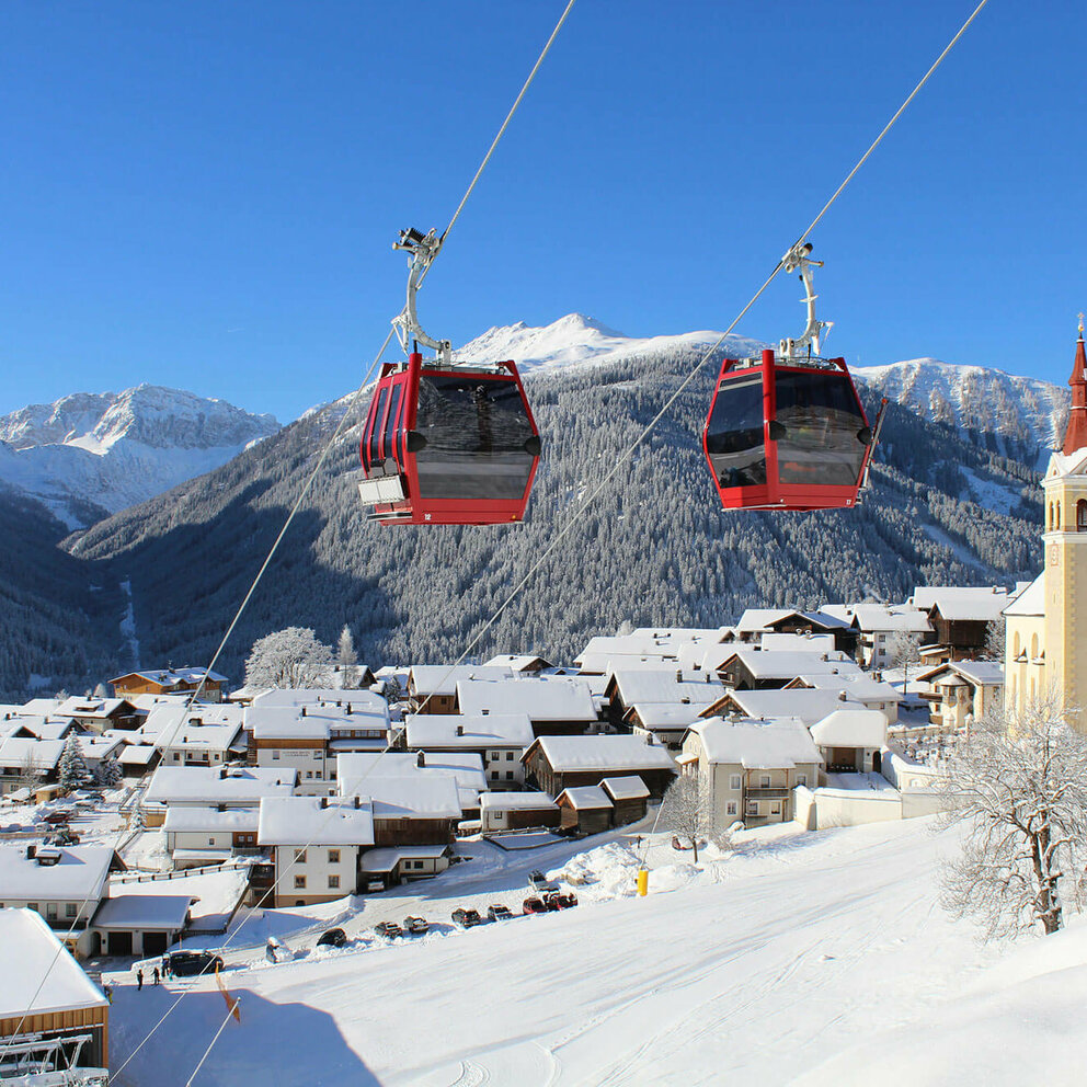 Zwei knallrote Gondeln der Bergbahn bilden einen herrlichen Kontrast zur tief verschneiten, sonnigen Winterlandschaft mit einem Haufendorf und großer Pfarrkirche am rechten Dorfrand. Tief blauer Himmel über dem verschneiten Bergkamm im Hintergrund.