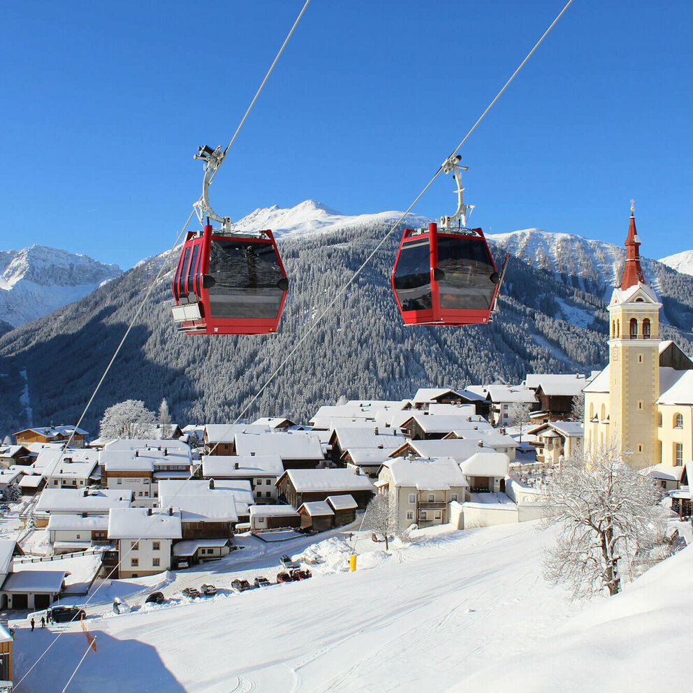 Zwei knallrote Gondeln der Bergbahn bilden einen herrlichen Kontrast zur tief verschneiten, sonnigen Winterlandschaft mit einem Haufendorf und großer Pfarrkirche am rechten Dorfrand. Tief blauer Himmel über dem verschneiten Bergkamm im Hintergrund.