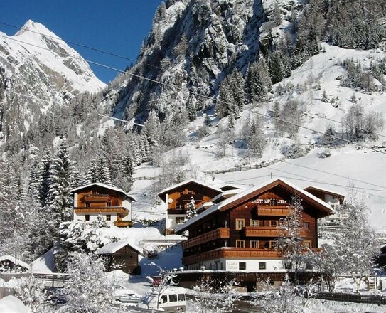 Das Bild zeigt den Gasthof Islitzer im Winter, umgeben von einer verschneiten Berglandschaft. Das mehrstöckige Gebäude in traditioneller Holzbauweise hebt sich mit seinen Balkonen und der dunklen Fassade deutlich von der weißen Schneedecke ab. Im Hintergrund sind steile, schneebedeckte Berghänge und zahlreiche Bäume zu sehen.