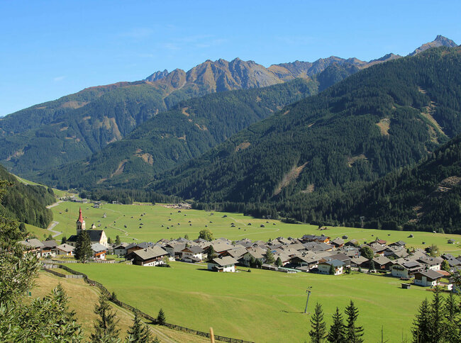 Blick auf eng verbautes Haufendorf mit ausschließlich grauer Dacheindeckung an einem wolkenlosen Herbsttag. Im Hintergrund die grünen Felder mit vielen kleinen Heuhütten und ein Gebirgszug, der das Tal mit seinen bewaldeten Hängen verschließt.