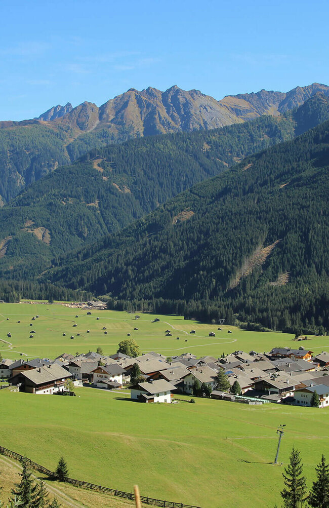 Blick auf eng verbautes Haufendorf mit ausschließlich grauer Dacheindeckung an einem wolkenlosen Herbsttag. Im Hintergrund die grünen Felder mit vielen kleinen Heuhütten und ein Gebirgszug, der das Tal mit seinen bewaldeten Hängen verschließt.