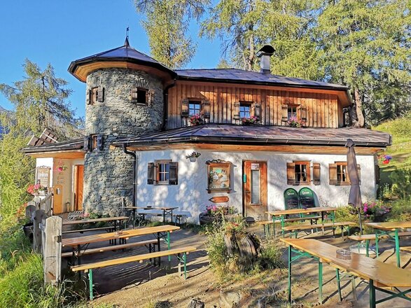 Das Bild zeigt die Stabanthütte, ein zweigeschossiges Gebäude mit heller Fassade, Natursteinmauerwerk und Holzverkleidung im oberen Bereich. Vor der Hütte befinden sich mehrere Holztische und Sitzbänke für Gäste. Die Hütte ist von Wiesen und Bäumen umgeben und fügt sich harmonisch in die Berglandschaft ein.