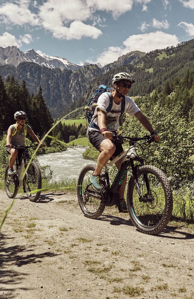 Zwei E-Bike Radahrerinnen fahren auf einem Schotterweg direkt am Fluss entlang. 